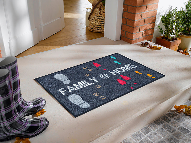 graue Fußmatte mit Schuhabdrücken und Schriftzug "FAMILY @ HOME" vor der Tür