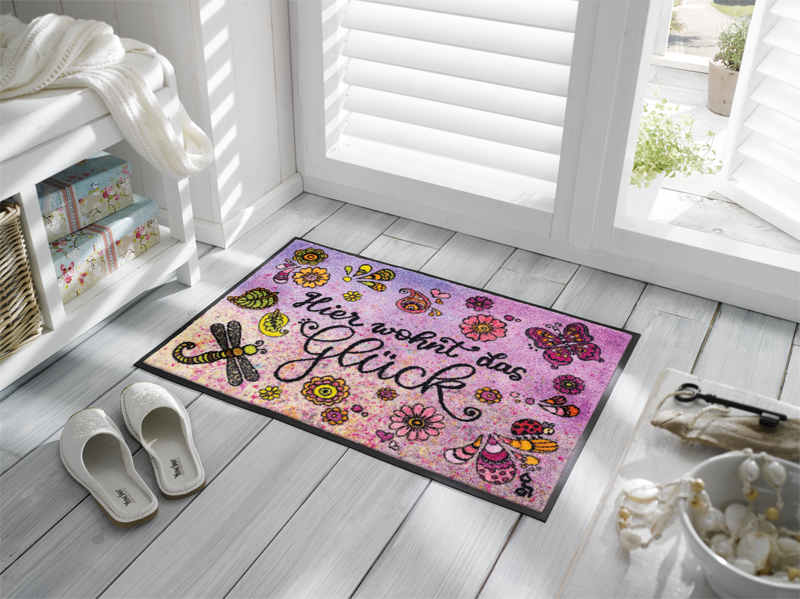 Fußmatte mit Schriftzug "Hier wohnt das Glück" und gemalten Blumen-Tiermotiven vor der Terrassentür